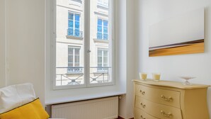 Une pièce avec un radiateur blanc, un canapé blanc orné d’un coussin jaune, une commode de couleur crème dotée de tiroirs, et une fenêtre donnant sur un immeuble avec des balcons.