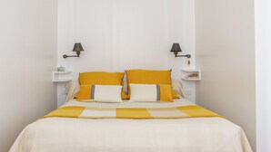 Un lit bien fait, recouvert d’une couvre-lit blanche et jaune, deux tables de chevet avec des lampes, et un mur blanc agrémenté d’un détail décoratif au niveau du plafond.