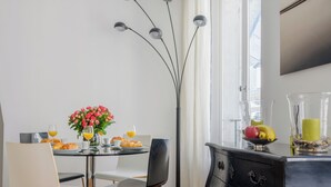 Un coin repas avec une table dressée pour deux personnes, un vase de fleurs et un panier de fruits posé sur un meuble noir.