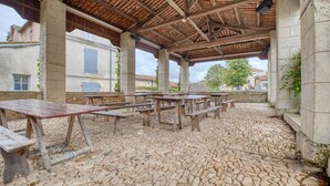 An outdoor covered patio with stone flooring, wooden tables and benches, and a view of buildings in the background.