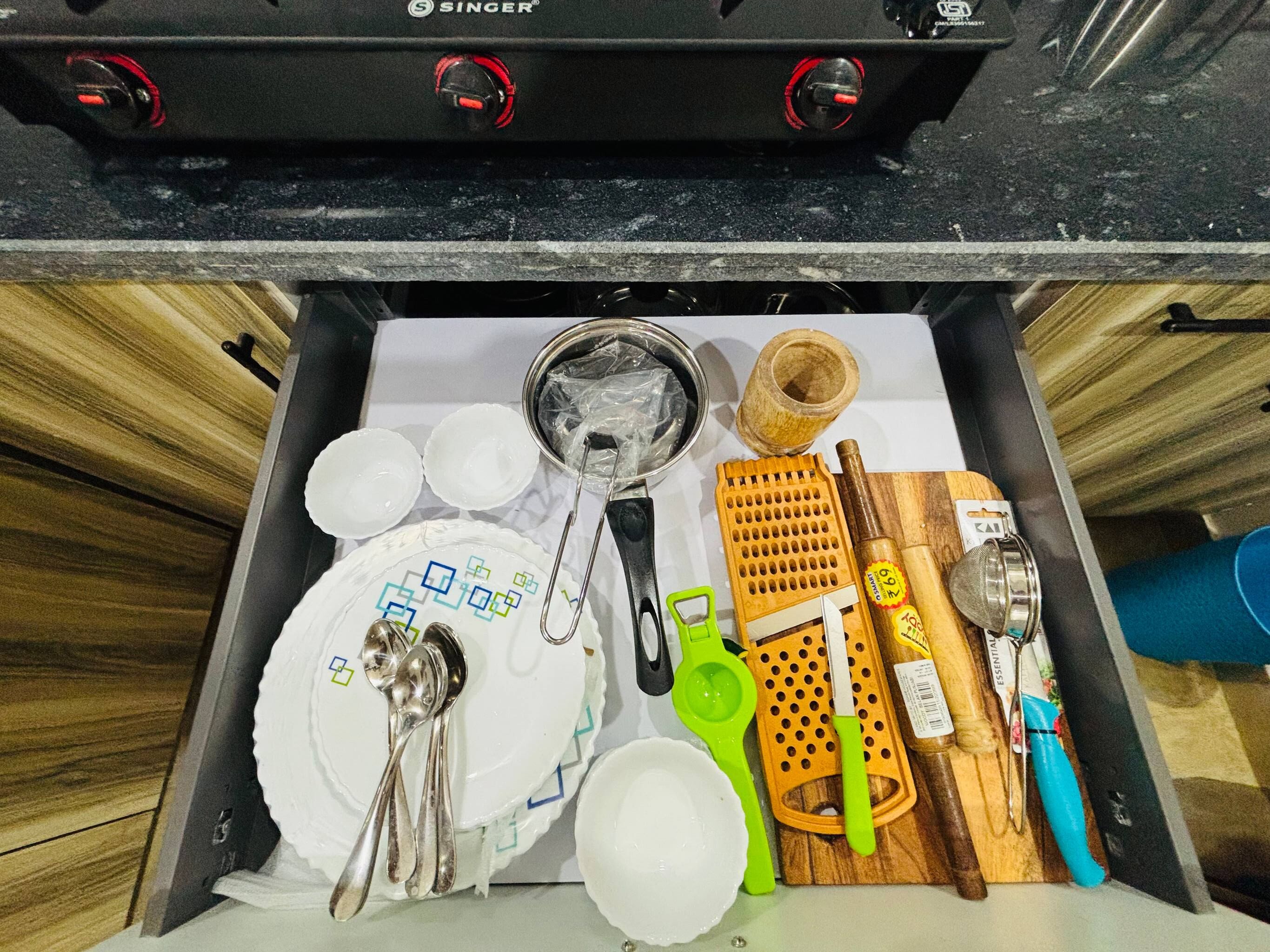 A drawer with kitchen utensils, including a grater, a bottle, and a plate with a spoon set.