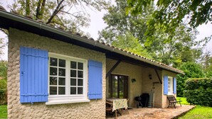 Une maison de campagne en pierre, avec des volets bleus, une véranda dotée d’une table et de chaises, et un barbecue à l’extérieur.