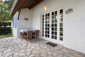 A covered patio with a stone floor, a table with chairs, and a white door with glass panels. - Gîte Le Lavoir - Domaine l'Étang des Mirandes with Shared Pool (Varaignes)