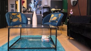 A modern living room with a glass coffee table, two blue armchairs with yellow cushions, a white vase with blue foliage, and a black sofa.