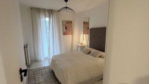 A bedroom with a bed, a nightstand, a lamp, and a window with curtains. - La Perle - T2 with terrace, air conditioning & parking near Toulouse city center (Toulouse)