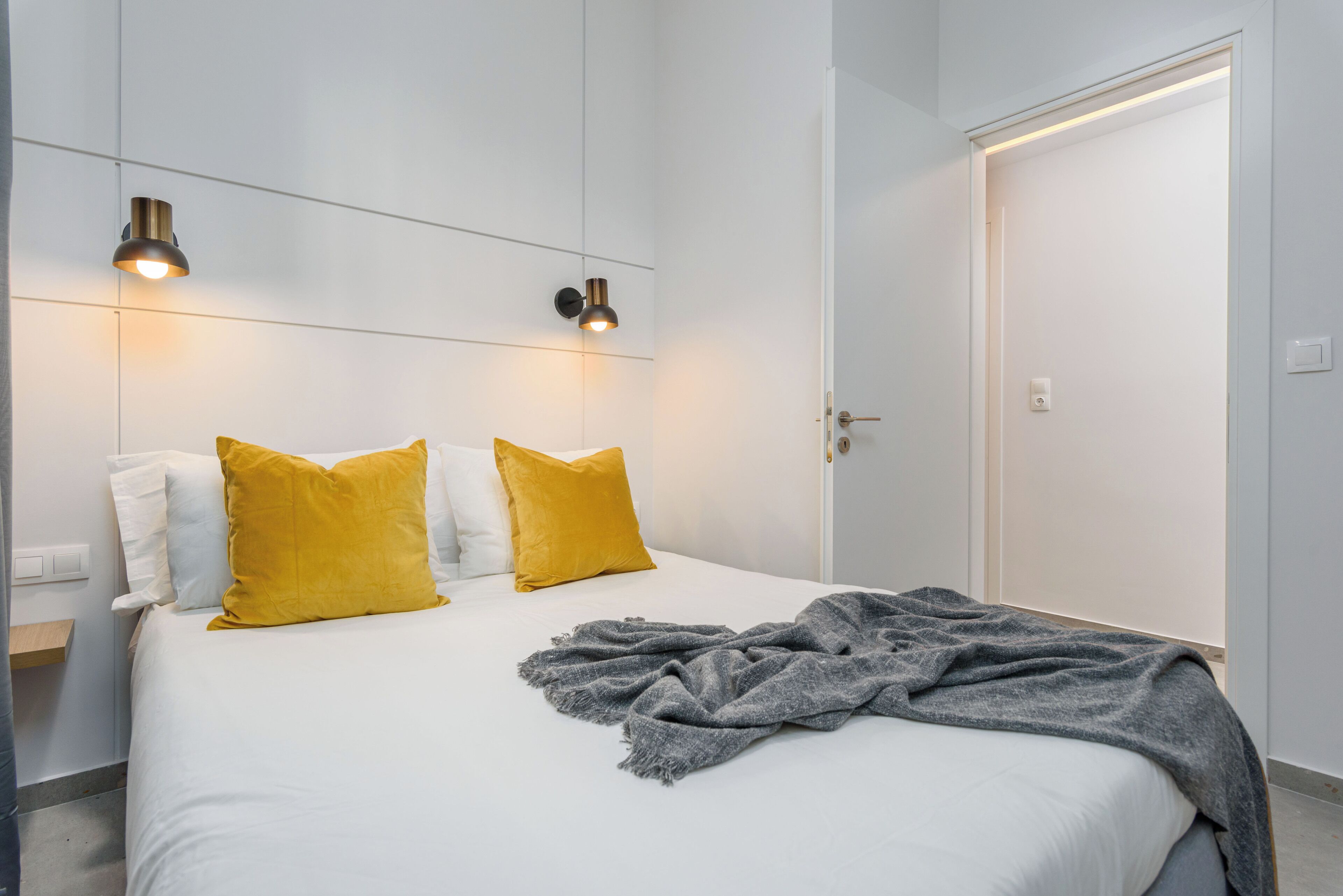 Un lit bien fait, agrémenté d’oreillers blancs et moutarde, d’un plaid gris et de deux lampes de lecture fixées au mur.
