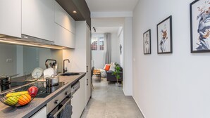 A modern kitchen with a built-in oven, a sink, and a countertop with a fruit bowl. There is a dining area with a table and chairs, and a living room with a sofa and a plant. - Penelope's Charming 2-BR Apt in Athens (Athens)