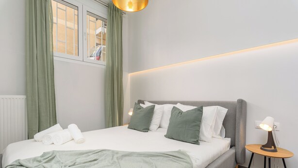 A neatly made bed with pillows and a blanket, a bedside lamp, and a view of the outside through a window with curtains. - Penelope's Charming 2-BR Apt in Athens (Athens)