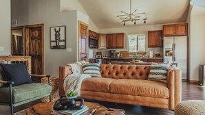 A spacious living room with a large brown sofa, a green armchair, and a wooden coffee table. The kitchen in the background features wooden cabinets and a stainless steel refrigerator.