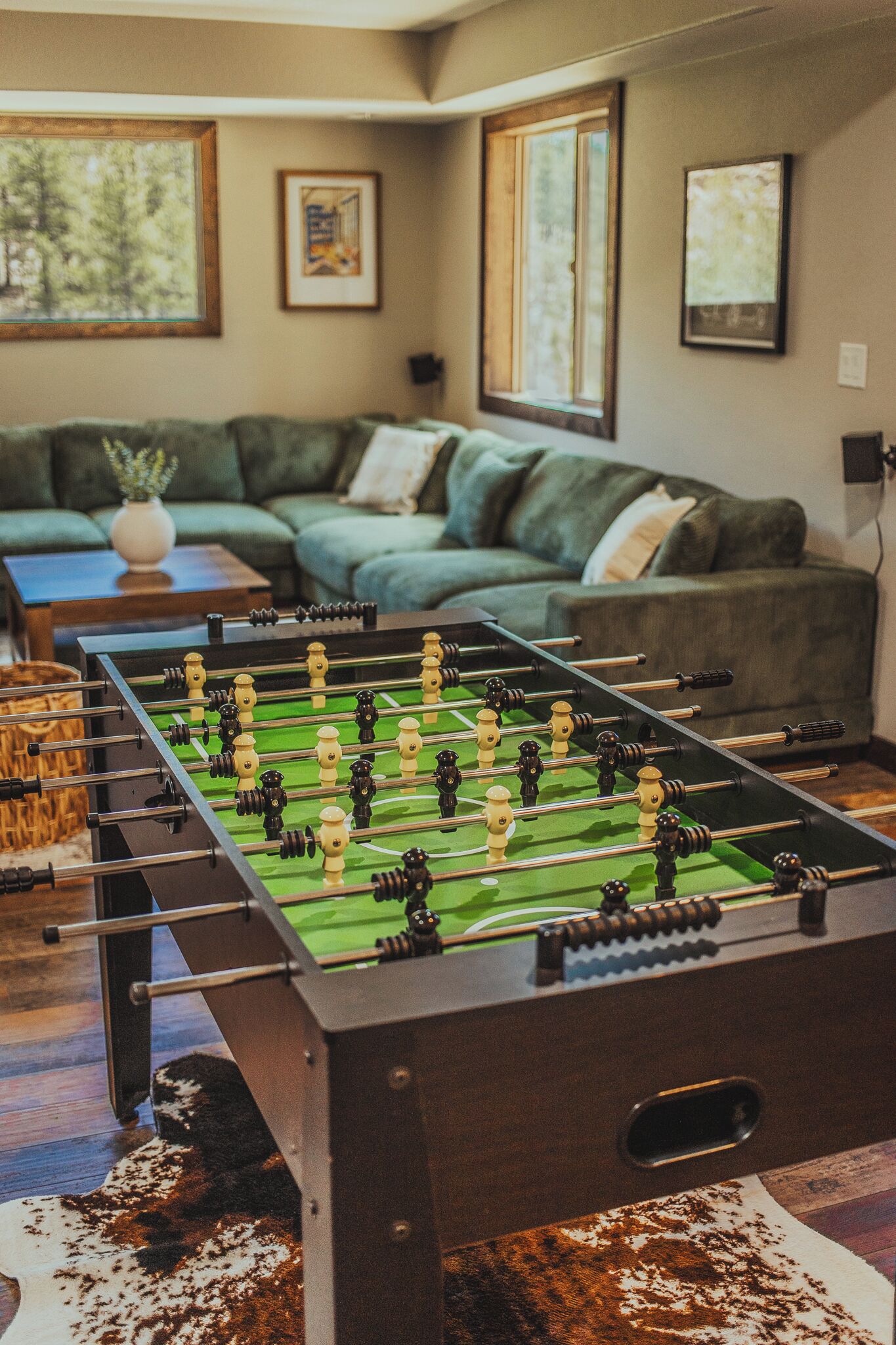 Une table de baby-foot dans une pièce avec un canapé vert, des tableaux encadrés au mur et une fenêtre.
