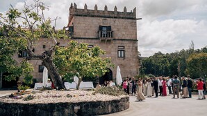 Un edificio histórico de piedra con una torre, rodeado de un jardín con mesas y sillas blancas.