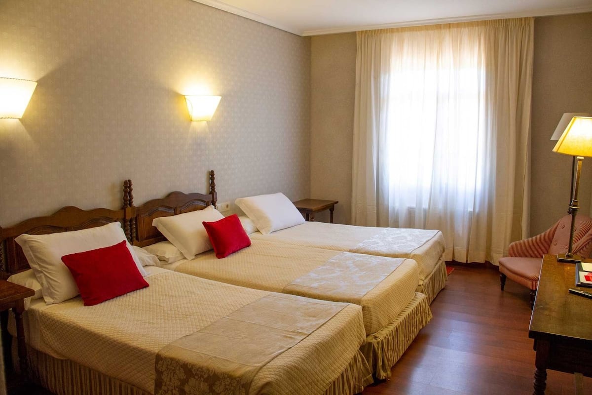 A hotel room with two beds, a wooden headboard, a small table, a chair, and a window with curtains.