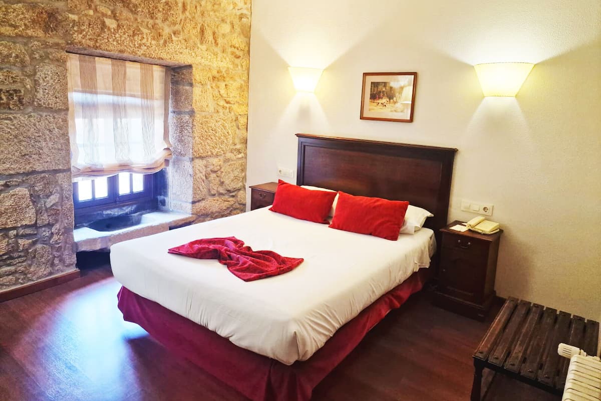 A bedroom with a stone wall, a bed with red and white bedding, a nightstand with a phone, and a window with curtains.