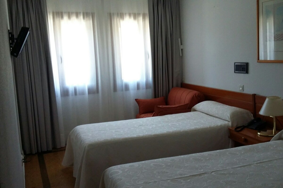 Habitación de hotel con dos camas, una mesita de noche, una lámpara y una ventana con cortinas.