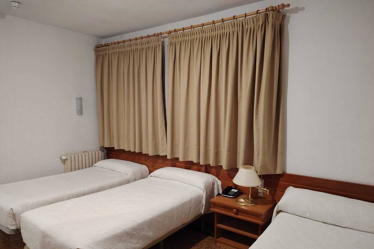 Habitación de hotel con dos camas individuales, una mesita de noche de madera con lámpara, teléfono y ventana con cortinas beige.