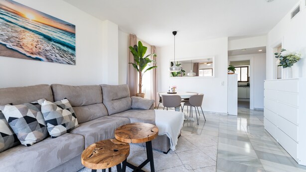 A modern living room with a sectional sofa, two wooden coffee tables, a dining area with a white table and chairs, and a kitchen in the background.