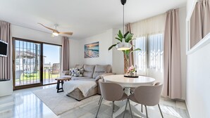 A modern living room with a sofa, a dining table with chairs, a ceiling fan, and a large window with curtains.