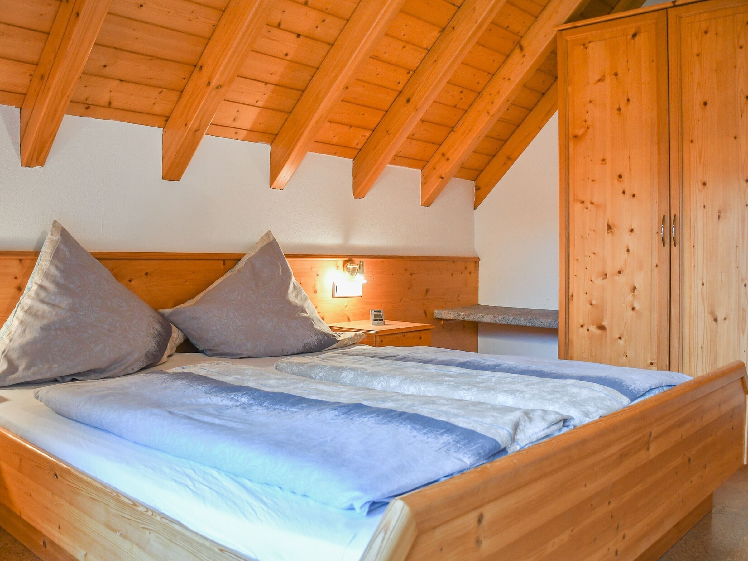 Een houten bed met kussens, een nachtkastje en een houten garderobe in een kamer met een schuine kap.