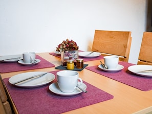 Una mesa de comedor dispuesta con platos blancos, tazas y cubiertos, junto con un jarrón con flores y un frasco pequeño, probablemente con salsas, sobre una mesa de madera con individuales morados.