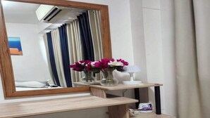 Une pièce comprenant un bureau en bois, un miroir, un petit tabouret, un vase avec des fleurs et une lampe.