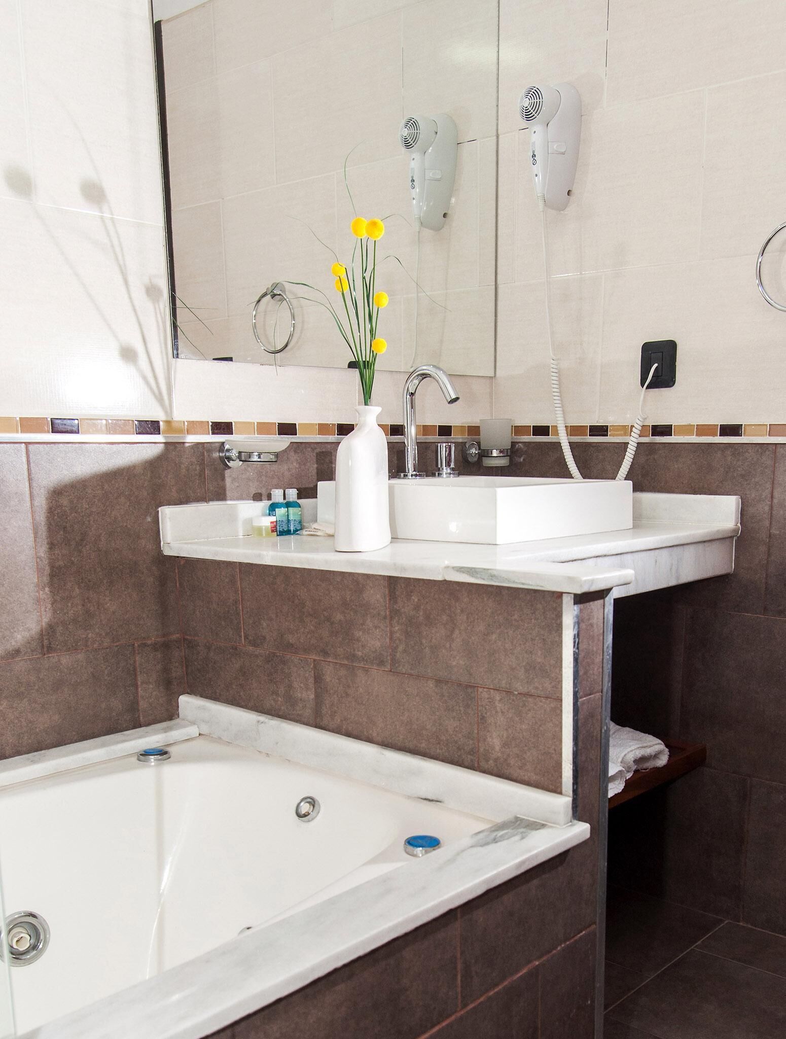 A bathroom with a white bathtub, two sinks, a mirror, a hairdryer, and a vase with yellow flowers.