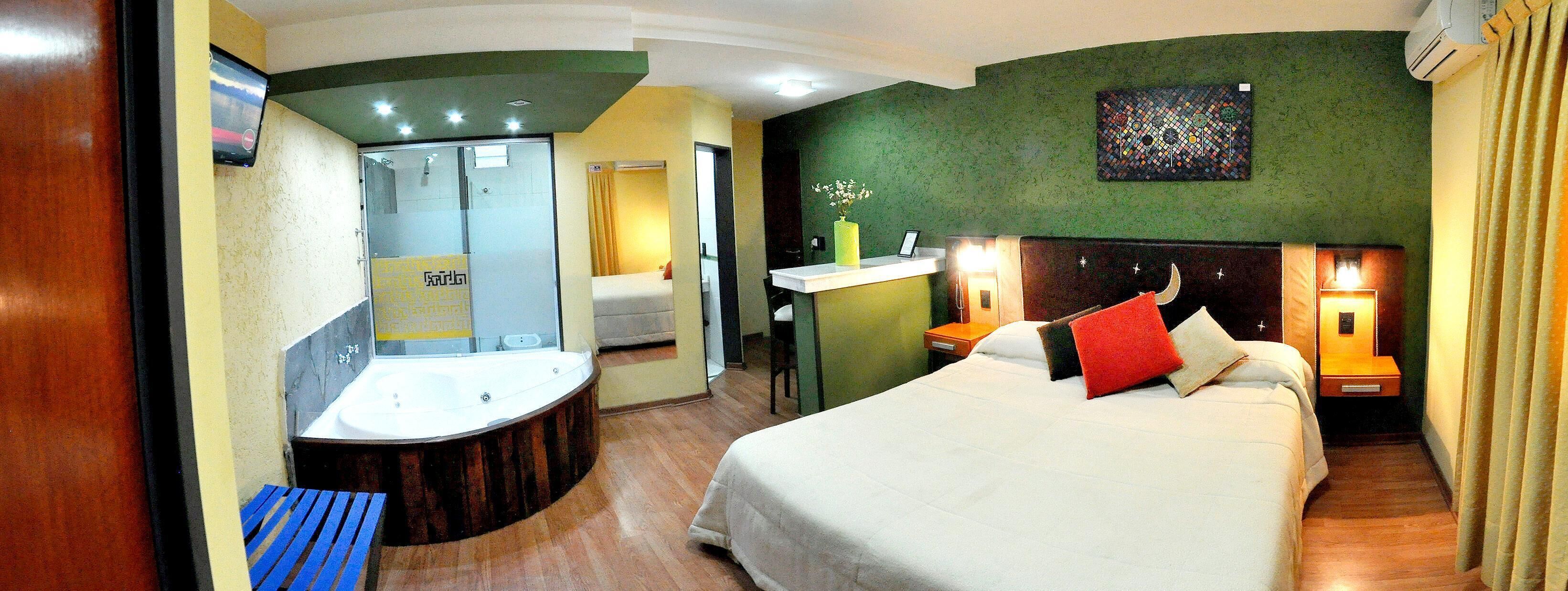 A hotel room with a bed, bedside tables, a nightstand, a mirror, a bathroom with a bathtub, and a wall-mounted air conditioner.