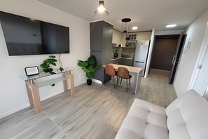 Ett modernt vardagsrum med en soffa, ett soffbord i glas och en tv som är monterad på väggen. Det finns också en matplats med ett bord och stolar, och ett kök längre in i rummet.