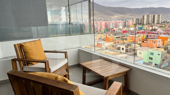 A balcony with a wooden chair and table, offering a view of colorful buildings and mountains.