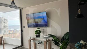 A modern living room with a large flat-screen TV mounted on the wall, a potted plant on a glass table, and a view of the cityscape through a large window.