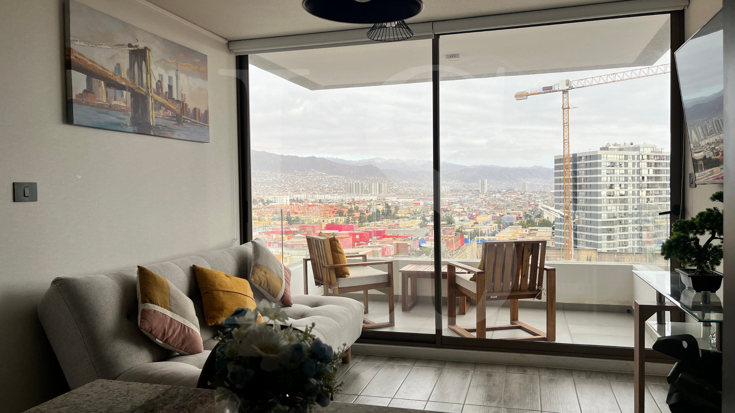 A modern living room with a sofa, a dining table, and chairs. A large window offers a view of a cityscape and mountains.