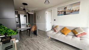 A modern living room with a grey sofa, a dining area with a table and chairs, and a kitchen in the background.