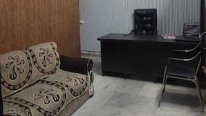 A room with a wooden wall, a sofa with a floral pattern, a desk with a chair, and a ceiling fan. - HOTEL STAY INN (Lucknow)