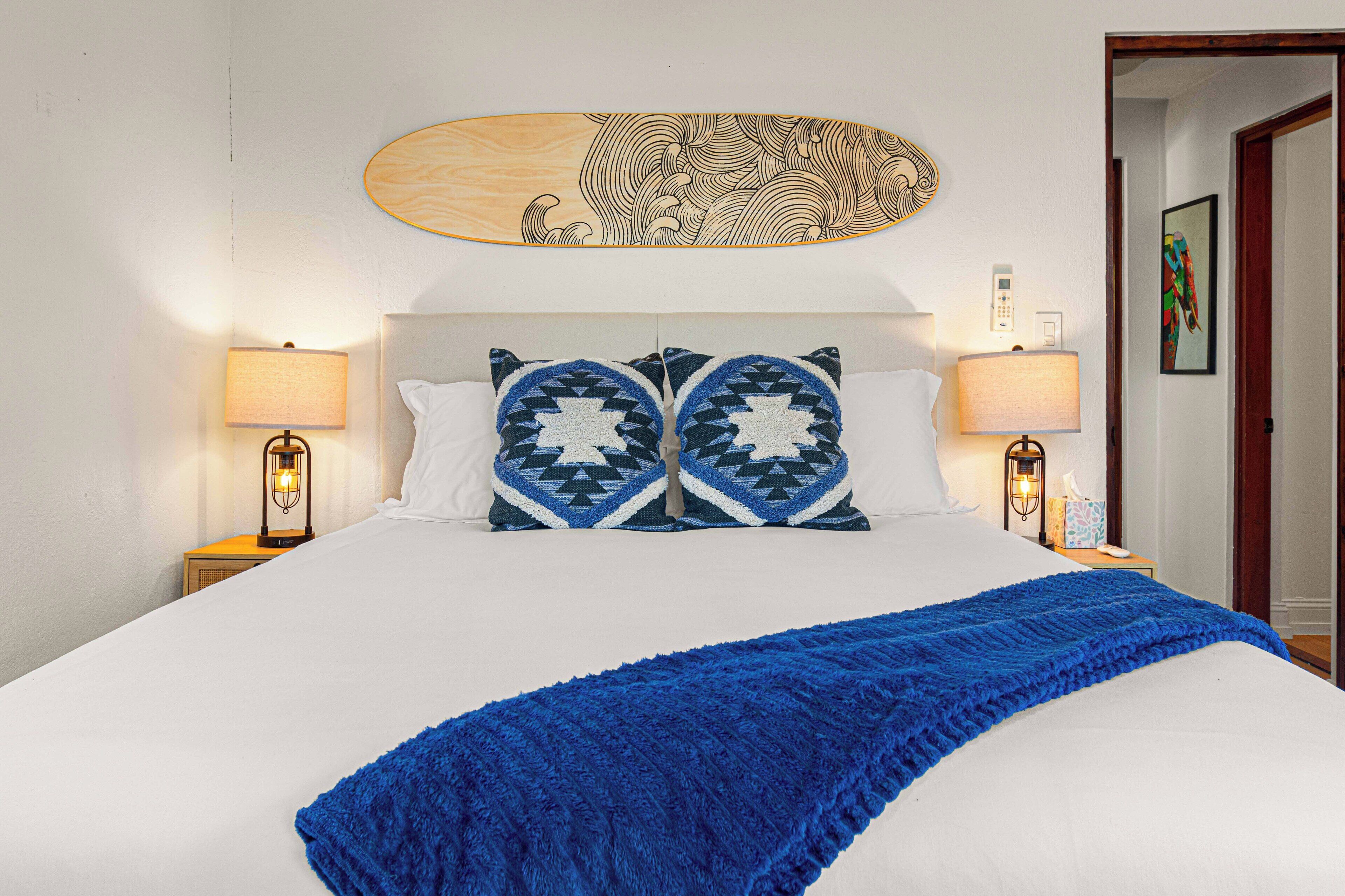 A neatly made bed with a blue blanket, two patterned pillows, and a wooden headboard with a decorative piece above.