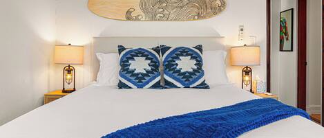 A neatly made bed with a blue blanket, two patterned pillows, and a wooden headboard with a decorative piece above.