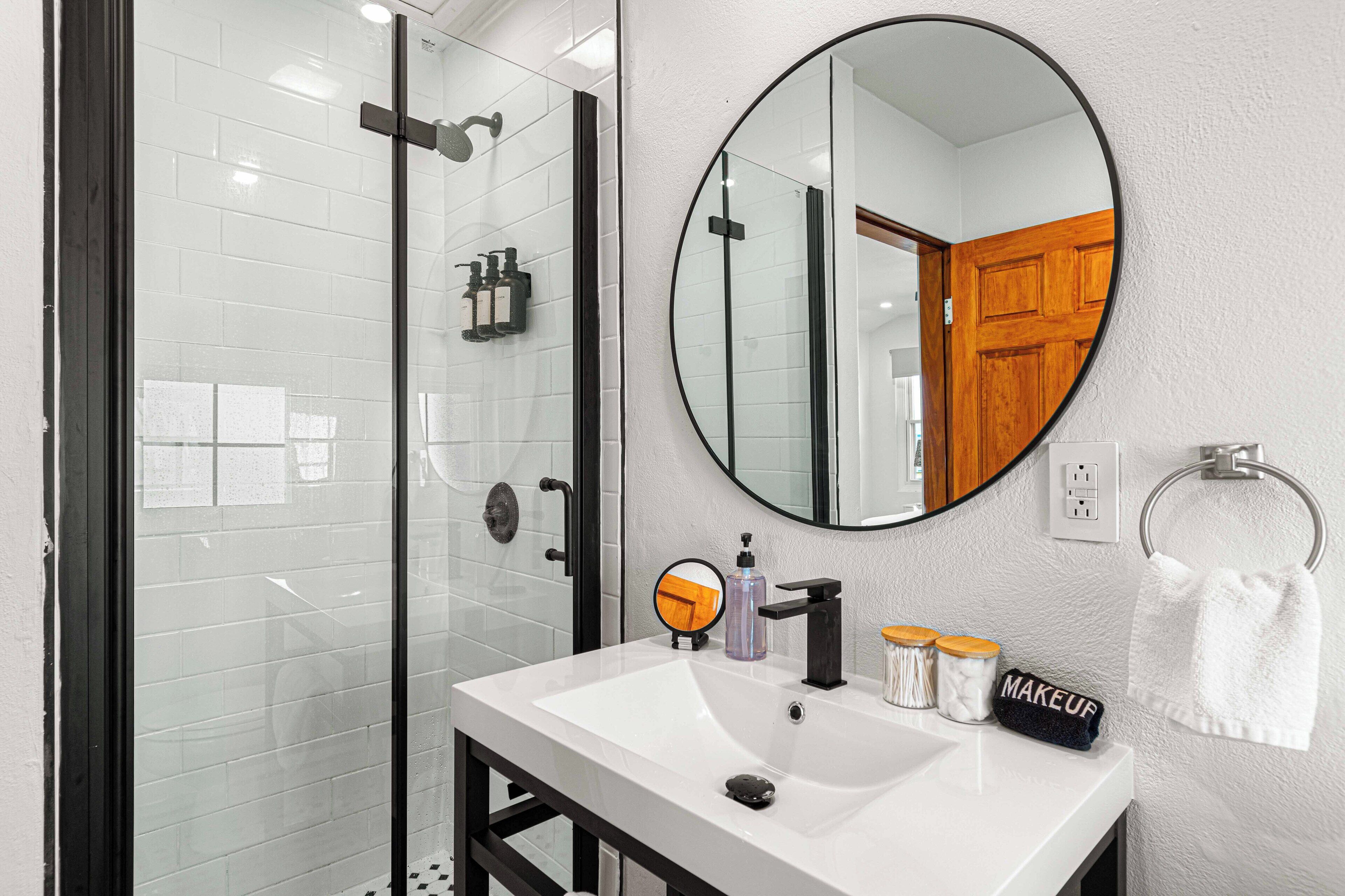 A bathroom with a glass-enclosed shower, a white sink with a black faucet, a round mirror, and a towel ring with a white towel.