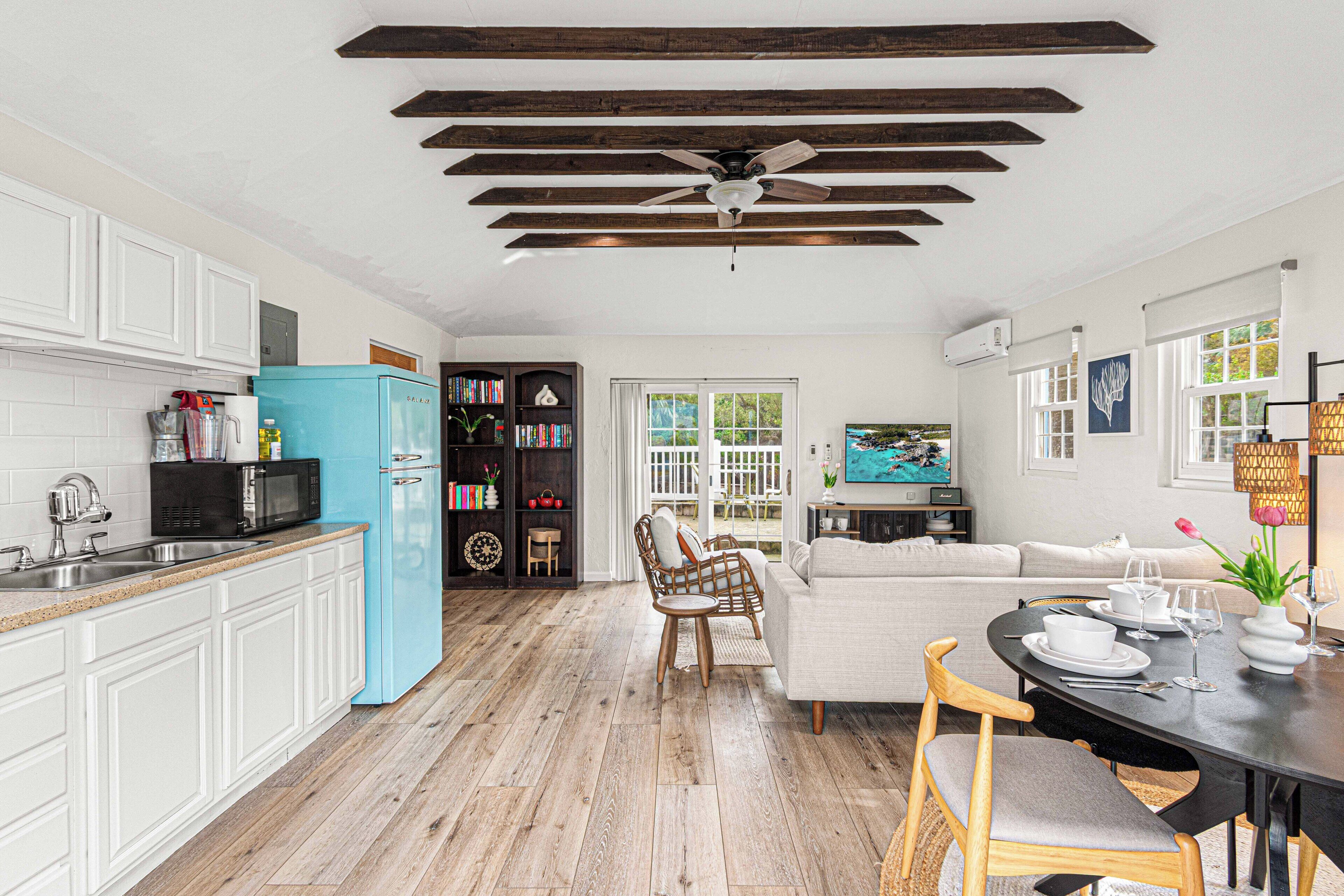 A spacious kitchen with white cabinets, a blue refrigerator, and wooden flooring. There is a dining area with a table set for four, a sofa, and a ceiling fan.