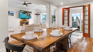 A dining area with a wooden table set for four, white plates, and wine glasses. There is a television mounted on the wall and a sofa in the background. - Blue Water (Southampton Parish)