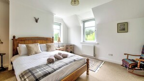 A bedroom with a large bed, a wooden headboard, a small side table, a rocking chair, and a window with a view of greenery. - Spacious Argyll Farmhouse in an Idyllic Setting (Dunoon)