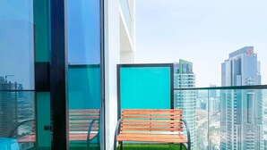 Un balcón con una silla de madera y un panel de vidrio, con vistas a un paisaje urbano con edificios modernos.