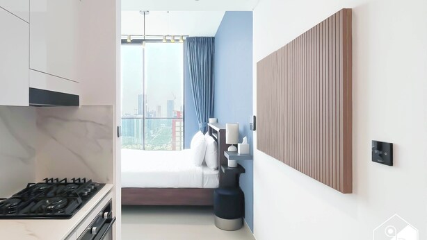 Habitación de hotel moderna con área de cocina, un amplio ventanal con vista a la ciudad y una mesita de noche con lámpara.