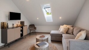 A modern loft bedroom with a sloped ceiling, a skylight, a flat-screen TV, a sofa, a chair, and a small round table.