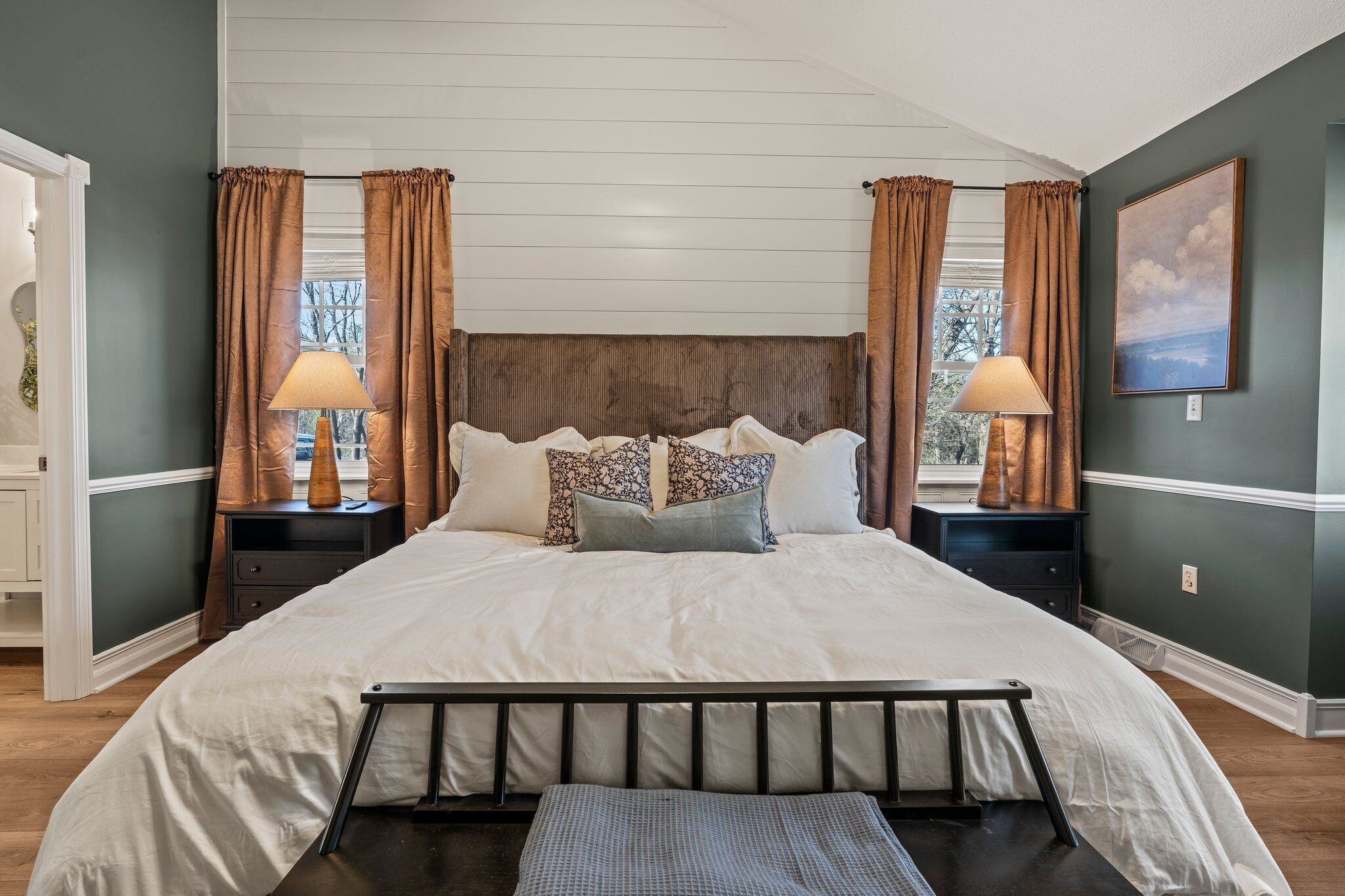 Une chambre avec un grand lit, deux tables de chevet, une fenêtre avec des rideaux et un tableau au mur.