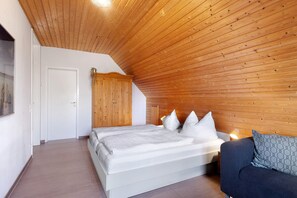 A bedroom with a wooden ceiling, a bed with white bedding, a nightstand with a lamp, and a sofa with patterned cushions.