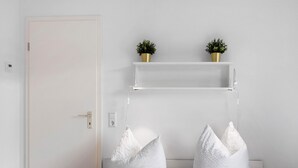 Une pièce avec une porte blanche, deux coussins blancs, une étagère blanche ornée de deux plantes en pot et une table de chevet blanche avec une horloge.