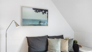 Une pièce moderne avec un canapé gris, une lampe sur pied, un bureau blanc et un tableau encadré représentant un banc au bord de la mer.