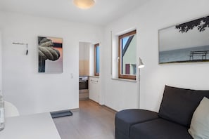 Un intérieur moderne avec un canapé, un tableau représentant une corde, un coin cuisine et une fenêtre donnant sur la mer.