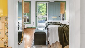 A bedroom with a bed, bedside table, and a mirror. There is a window with a view of greenery outside.