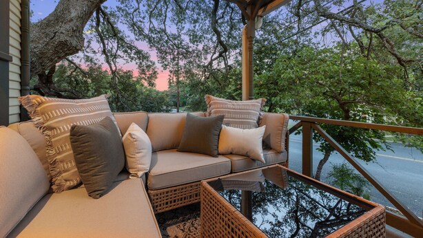 A covered outdoor seating area with a cushioned sofa and wicker furniture, overlooking a road and trees.