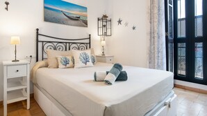 Una cama bien hecha con una colcha blanca y toallas azules, una mesita de noche con una lámpara, un cuadro colgado en la pared que muestra una playa, y una ventana con vistas al exterior.