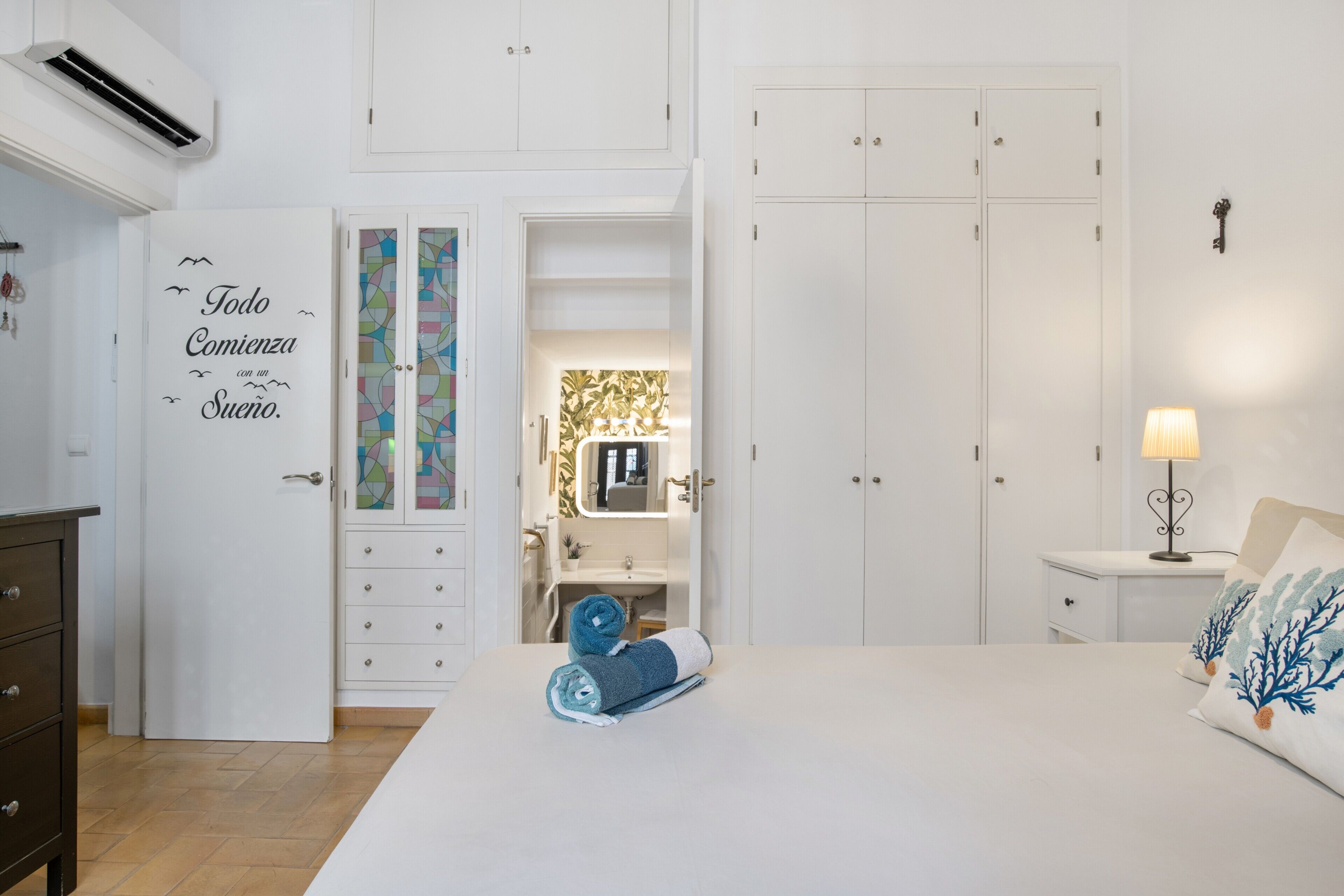A bedroom with a bed, a nightstand, a lamp, a dresser, a wardrobe, and a bathroom with a mirror and sink.